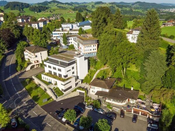 Moderne Klinik im Grünen mit mehreren Gebäuden, umgeben von Bäumen und Hügeln.