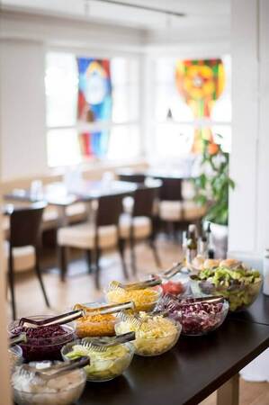 Vielfältiges Salatbuffet mit frischem Gemüse in hellem Restaurant mit bunten Fenstern.