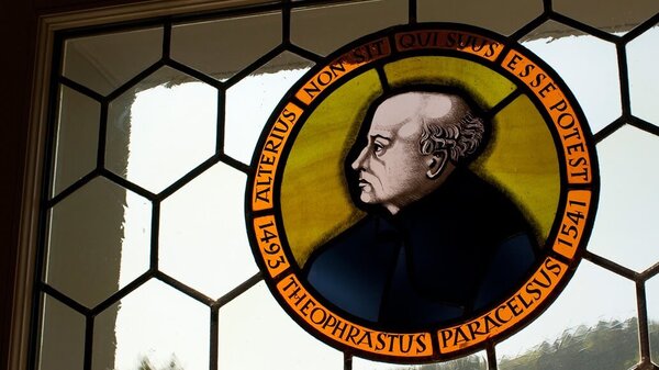 Buntglasfenster mit Porträt von Paracelsus und lateinischer Inschrift im runden Rahmen.